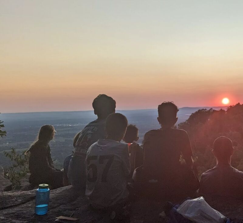 A group of people are sitting on a rocky outcrop, silhouetted against a vibrant sunset. The sky is a blend of orange, pink, and yellow hues, with the sun appearing as a bright orb on the horizon. The landscape below is a mix of trees and distant hills, partially obscured by the atmospheric haze. A water bottle sits in the foreground, adding a touch of everyday life to the scenic view.